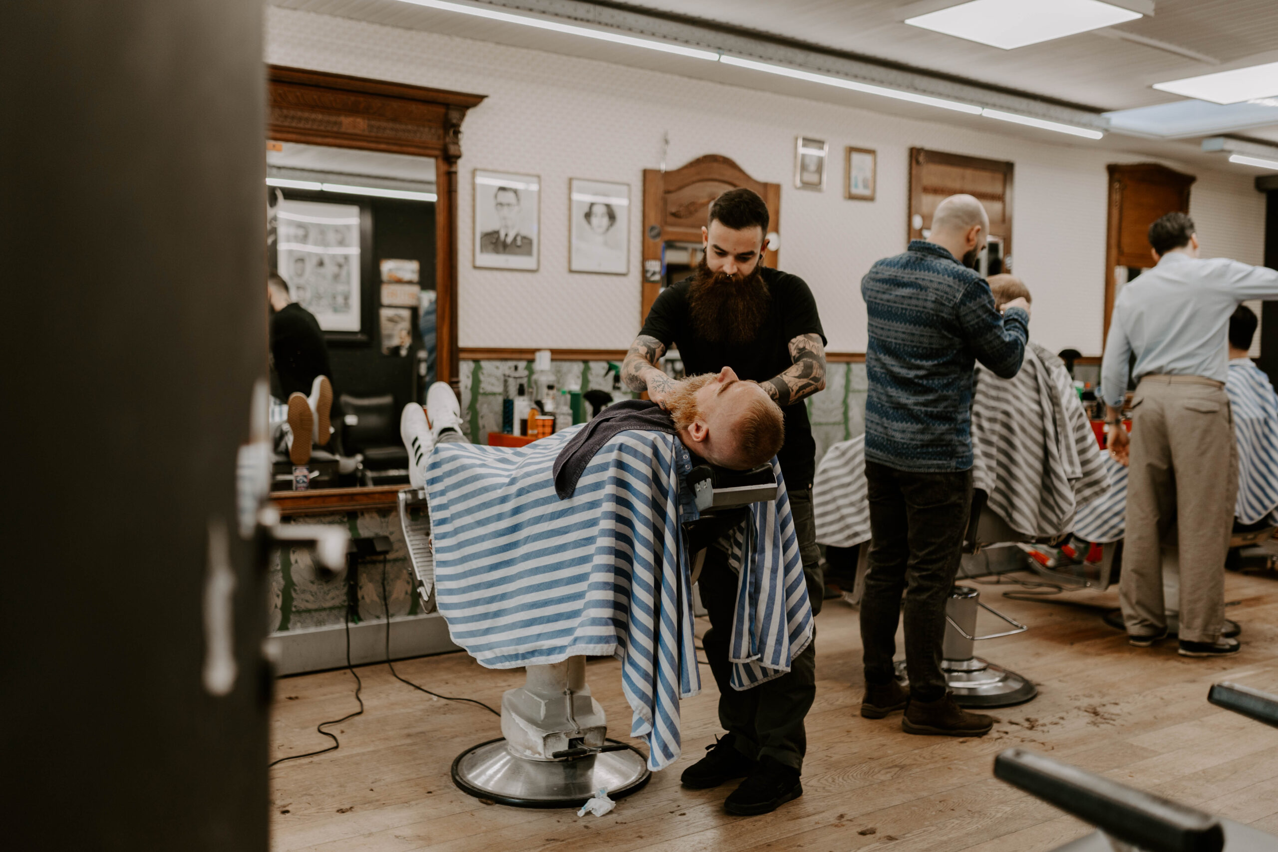 barbiers aan het werk. een open deur toont de binnenkant van een barbershop, waar 3 barbiers aan het werk zijn. Een eerste barbier is aan het scheren, twee andere barbiers zijn aan het knippen. Het ziet er gezellig druk uit. Het is een zeer uitnodigend beeld.
